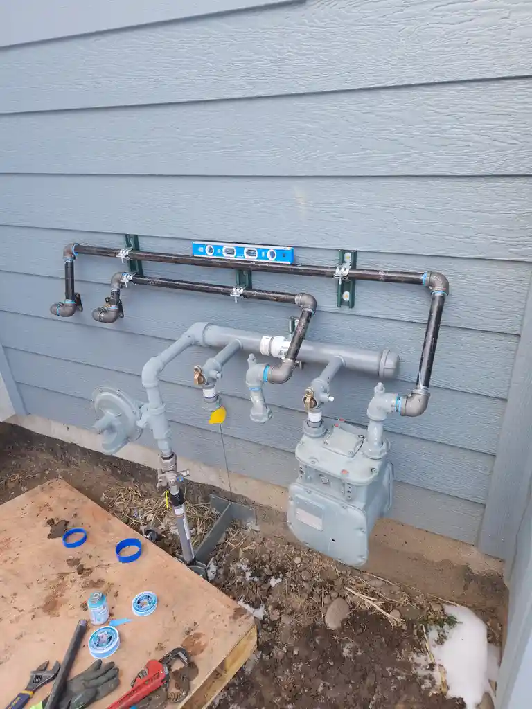 An outdoor gas line installation on a gray house wall features a blue spirit level placed horizontally across the top pipe…