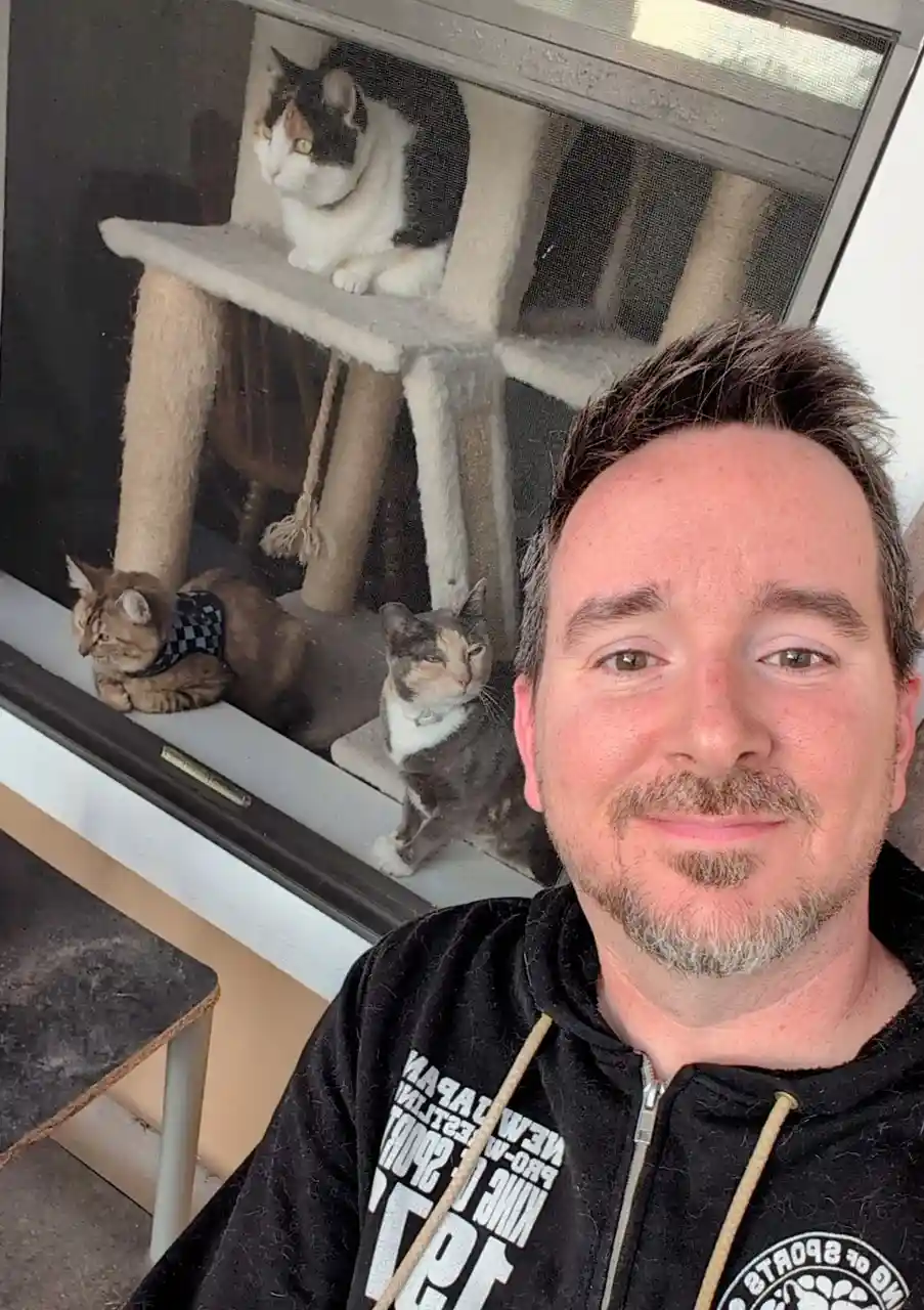 A man takes a selfie looking at the camera with a slight smile, positioned in front of a screen door. Behind him, three cats…