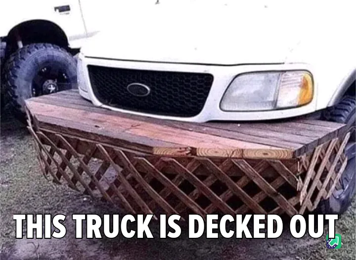 A white truck has a large wooden platform with lattice sides bolted to its front bumper, making it look like a deck. The…