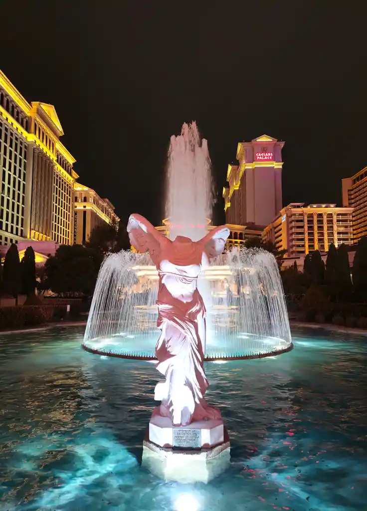 A nighttime photo of the iconic winged goddess statue at the Caesars Palace fountain in Las Vegas, with water cascading…
