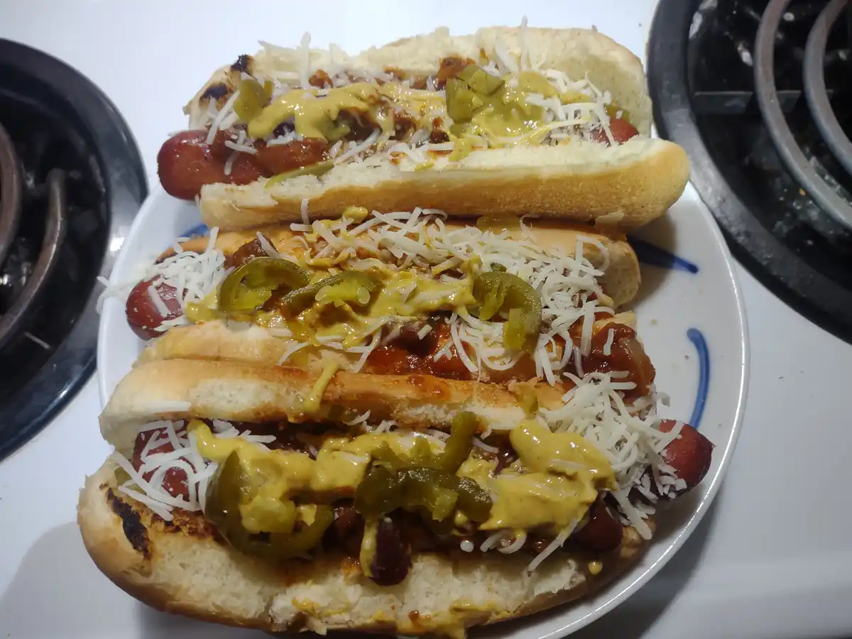 Three heavily loaded hot dogs on a white plate with blue trim, sitting on a white stovetop with black burners visible on…