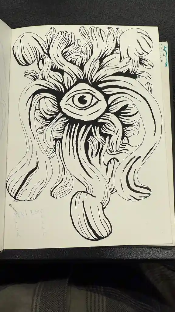 A black and white ink drawing in a sketchbook shows a surreal, organic creature with a large central eye surrounded by…