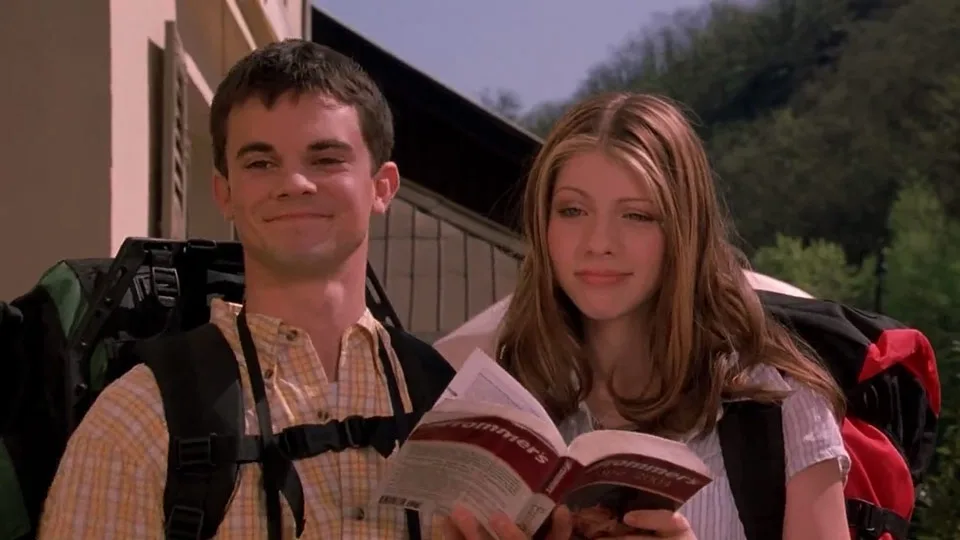 A man and woman, both wearing backpacks, stand outdoors holding an open travel guidebook titled 'Summer'. The man has a…