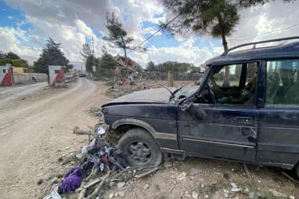 A dark blue, heavily damaged car sits on a dirt road with its front end crushed and debris scattered around. A motorcycle is…