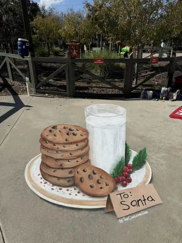A 3d chalk drawing on pavement depicts a plate with a tall stack of chocolate chip cookies, a glass of milk, holly berries…