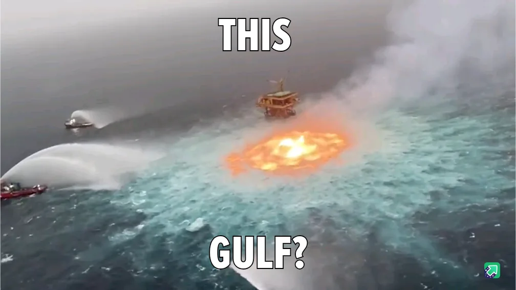 An aerial view shows a large offshore oil rig on fire in the ocean, with bright orange flames and thick smoke rising…