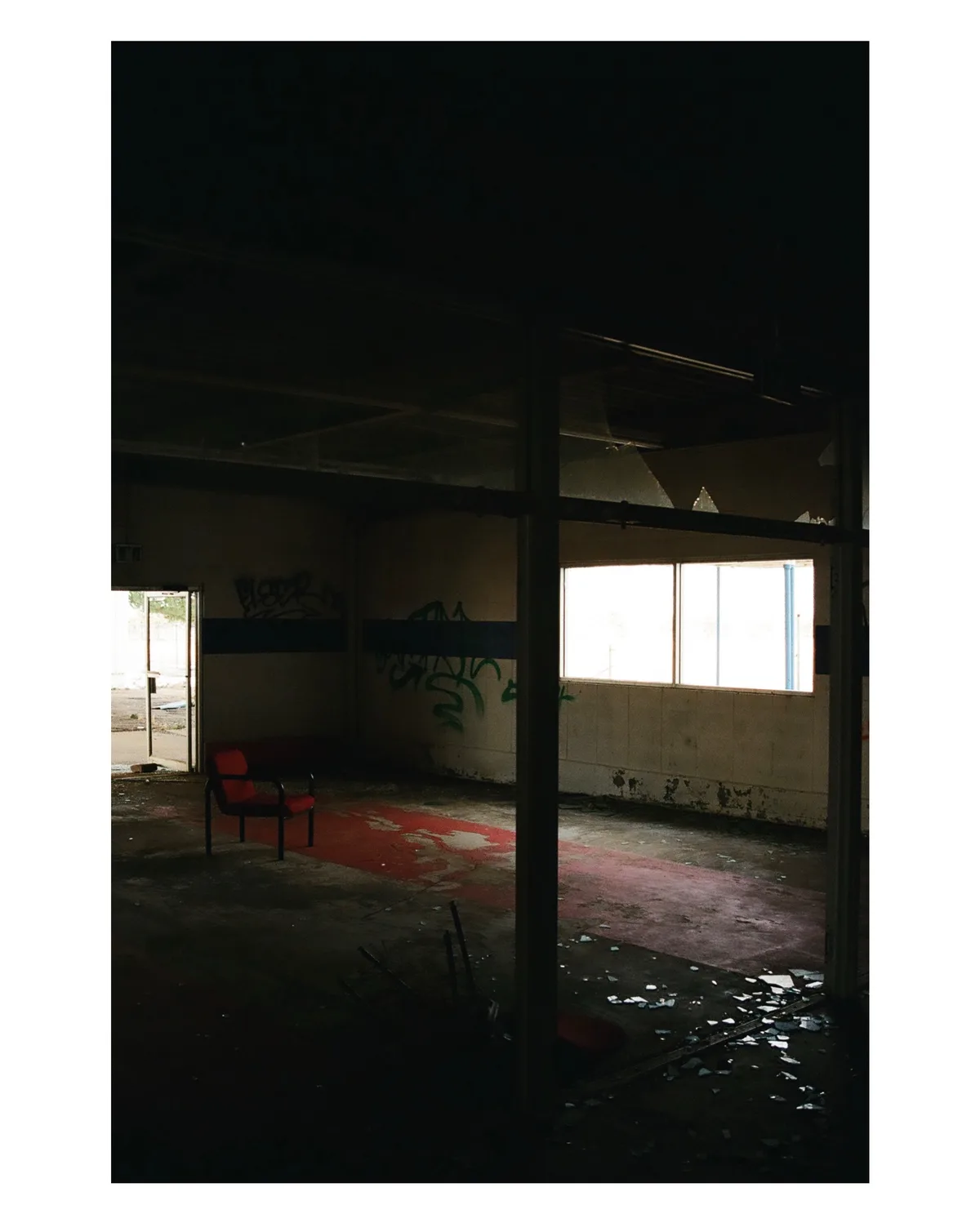 A dimly lit, abandoned indoor space with graffiti on walls, broken glass on the floor, and a single red chair in the…