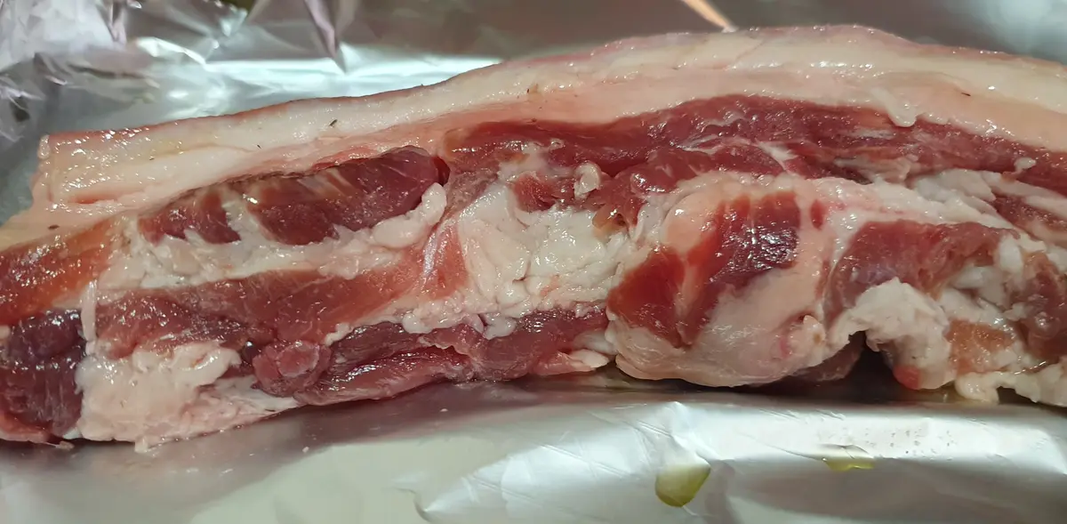 A close-up photo shows a raw rack of ribs with visible red meat and white fat layers resting on crinkled aluminum foil. A…