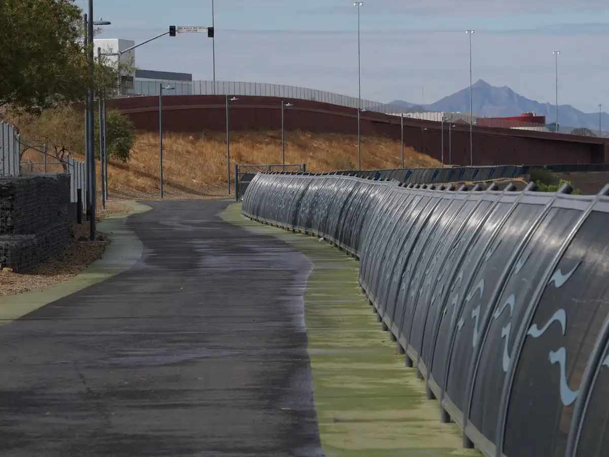 A wet paved path curves alongside a long, curved, dark gray metal fence with light blue wavy patterns, set against a dry…