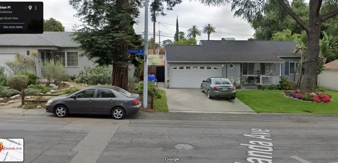 A google street view image of a suburban house with two cars parked, one in the driveway and one on the street. A person in…