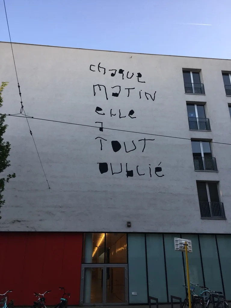 A plain white apartment building facade has large, black, handwritten-style graffiti that reads 'chaque matin elle TOUT…