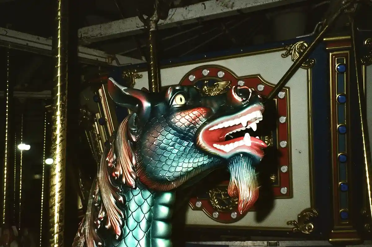 A close-up of a colorful, ornate carousel horse with a dragon-like head, featuring scales, sharp teeth, and a fierce…