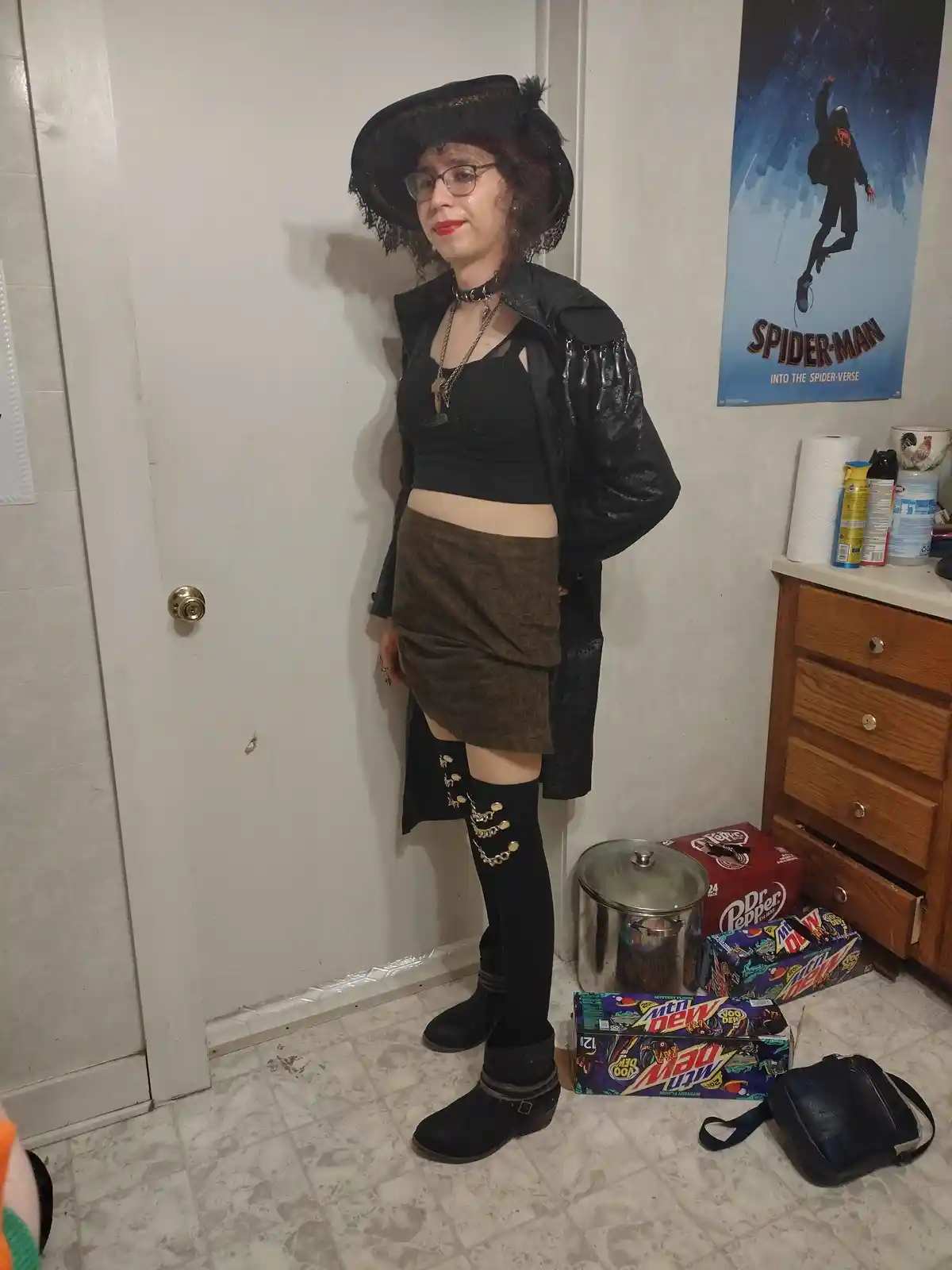 A person in a dark, gothic-style outfit with a wide-brimmed hat, glasses, and thigh-high boots stands in a bathroom. They…
