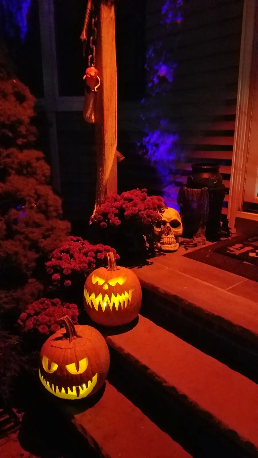 A nighttime photo of a Halloween-decorated porch with two glowing jack-o'-lanterns, a plastic skull, red flowers, and a…