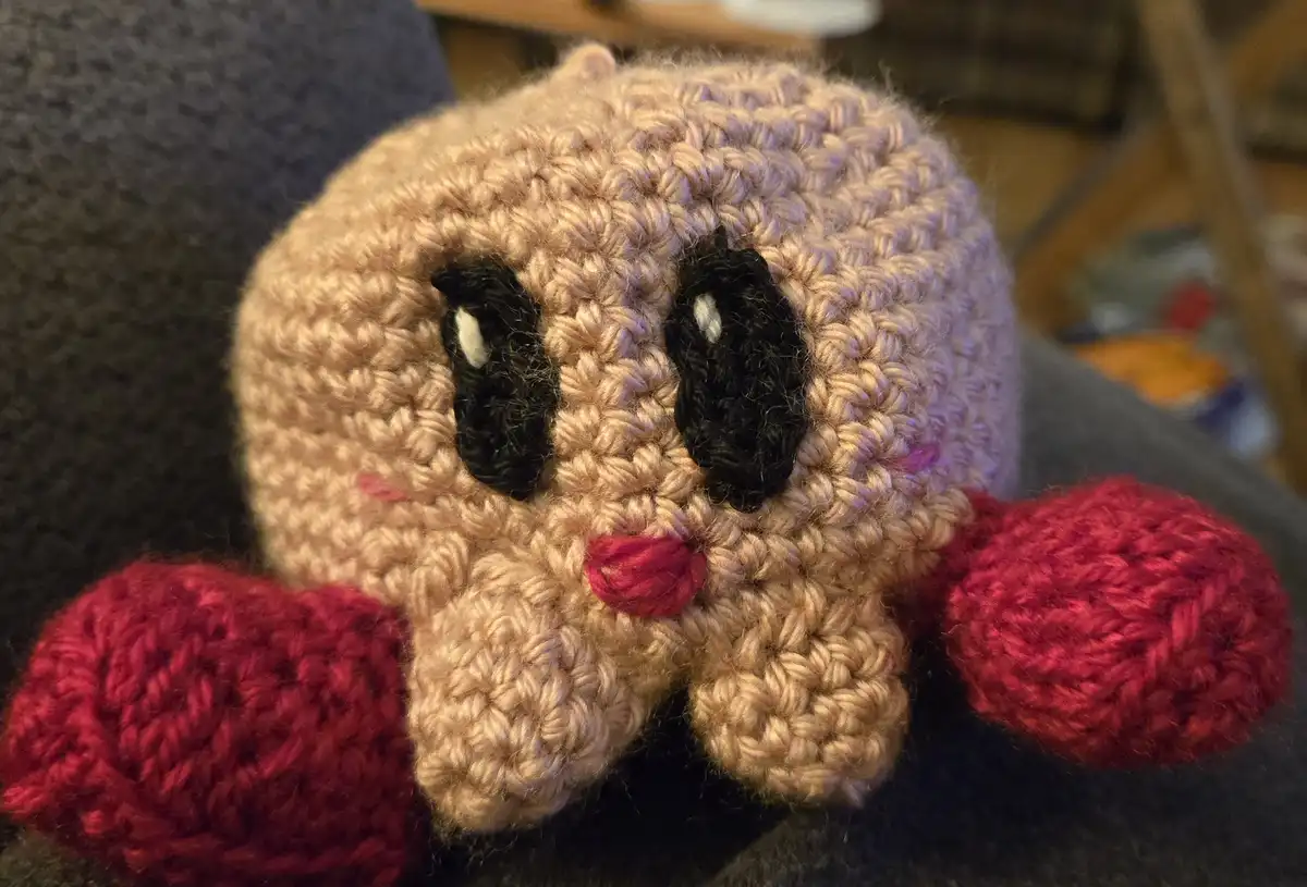 A close-up photo of a handmade, beige crocheted plush toy resembling a round, cute creature with black eyes, pink cheeks…