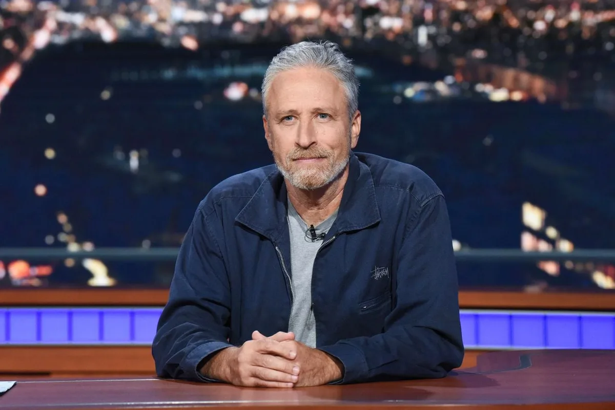 a man with gray hair and a beard, wearing a dark blue jacket over a gray shirt, sits at a desk with his hands clasped…
