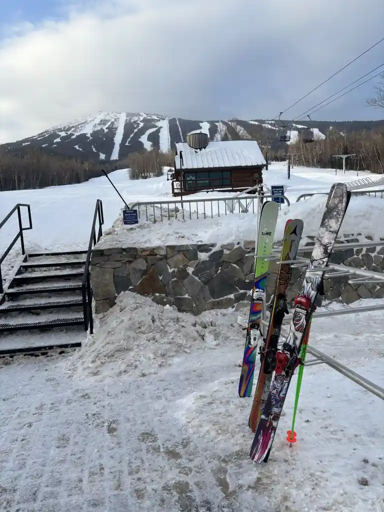 A snowy ski resort scene with three colorful skis leaning against a metal rack in the foreground. Behind them is a stone…