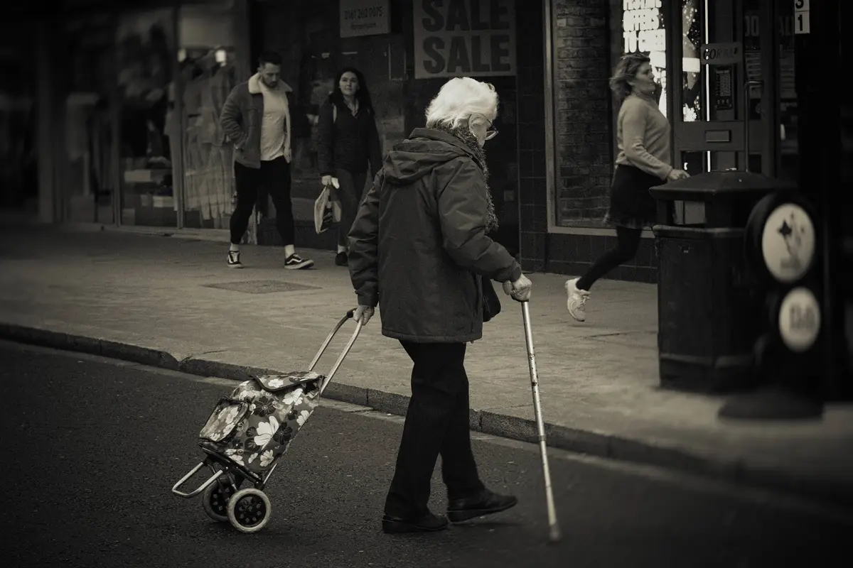 A black-and-white photo shows an elderly woman with white hair, wearing a dark coat and scarf, walking on a city street…