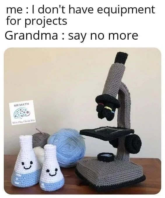 A crocheted microscope and two smiling crocheted beakers sit on a wooden table next to yarn and a small sign that reads 'MR…