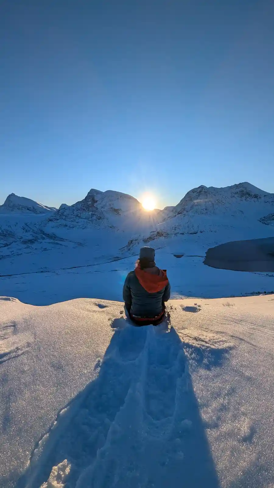 A person in winter gear sits on a snowy mountain ridge, facing a sunrise over snow-capped peaks, with their long shadow…