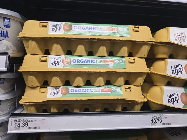 Stacks of yellow cardboard egg cartons labeled 'Happy Egg Organic Free Range 18 Eggs' sit on a supermarket shelf. A price…