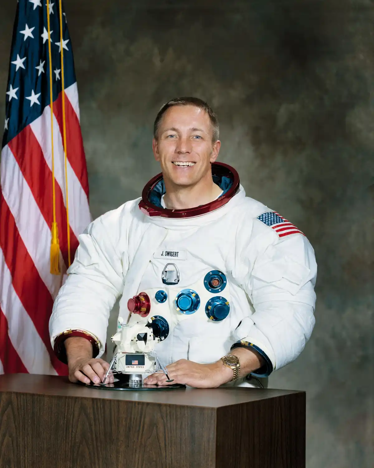 A smiling astronaut in a white spacesuit with 'J. SWIGERT' on his chest and an American flag patch on his sleeve, sits at a…