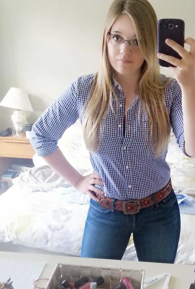 A young woman with glasses and long blonde hair takes a mirror selfie in a bedroom, wearing a blue and white checkered…