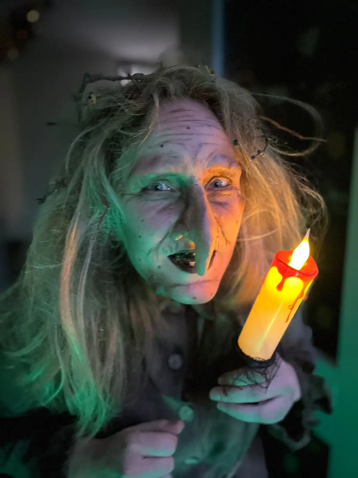 A person dressed as a witch with pale makeup, wild hair, and a twig crown holds a lit candle, their face illuminated by…