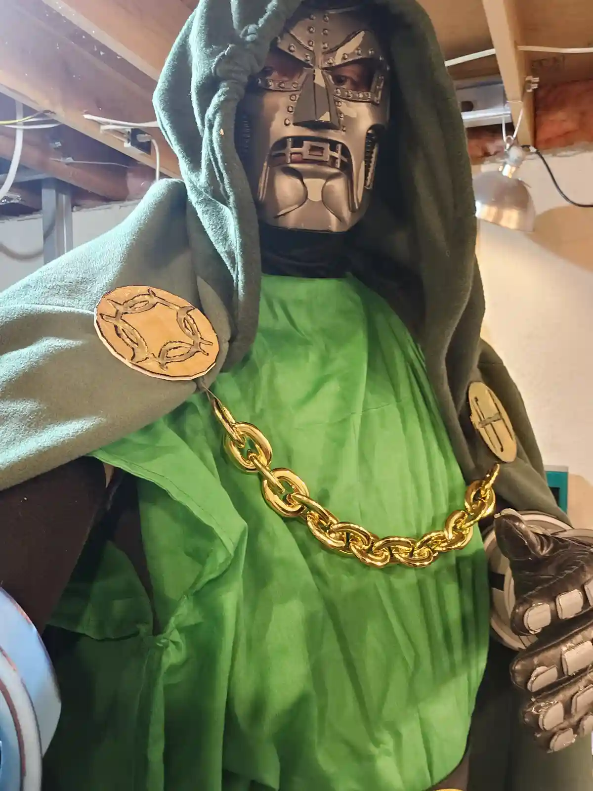 A person in a homemade Doctor Doom costume, featuring a metallic mask, green robe, and a large gold chain, stands in a room…
