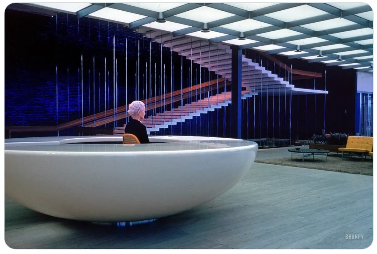 A woman with short white hair sits in a large, white, bowl-shaped desk in a futuristic office with a suspended staircase and…
