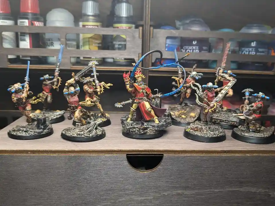 A row of painted miniature figurines, mostly in red and gold armor with futuristic weapons, are displayed on a wooden shelf…