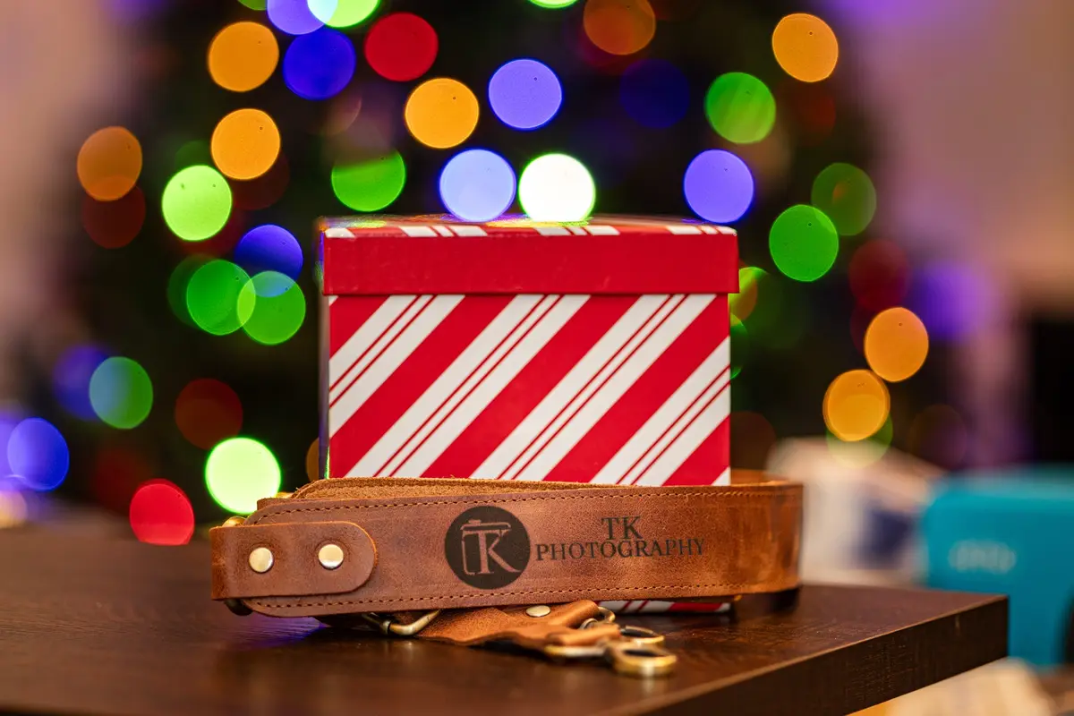 A brown leather camera strap with 'TK PHOTOGRAPHY' and a logo is draped over a red and white striped Christmas gift box. The…