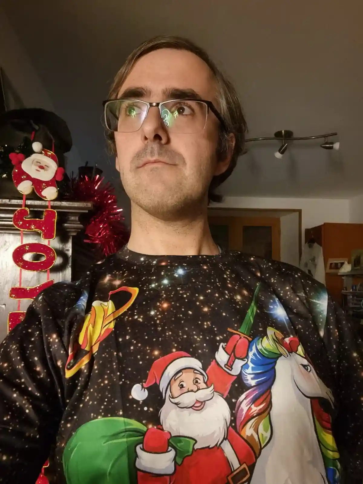A man wearing glasses and a galaxy-themed Christmas sweater featuring Santa Claus riding a rainbow unicorn looks upward with…