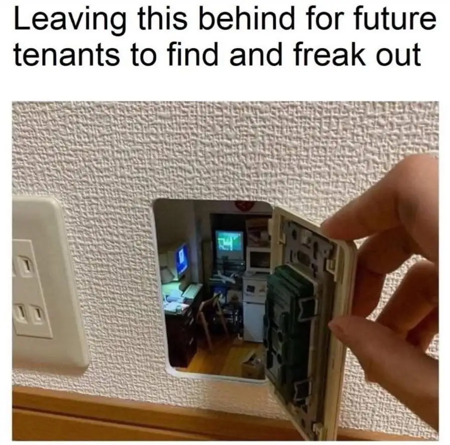 A hand holds open a wall plate, revealing a hidden compartment that looks into a cluttered room with multiple monitors and…