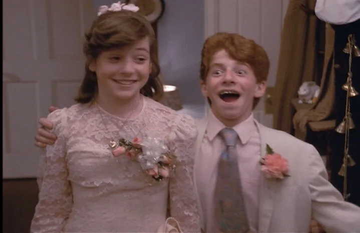 A still from a movie shows a young girl in a pink lace dress and a young boy in a light-colored suit, both wearing corsages…