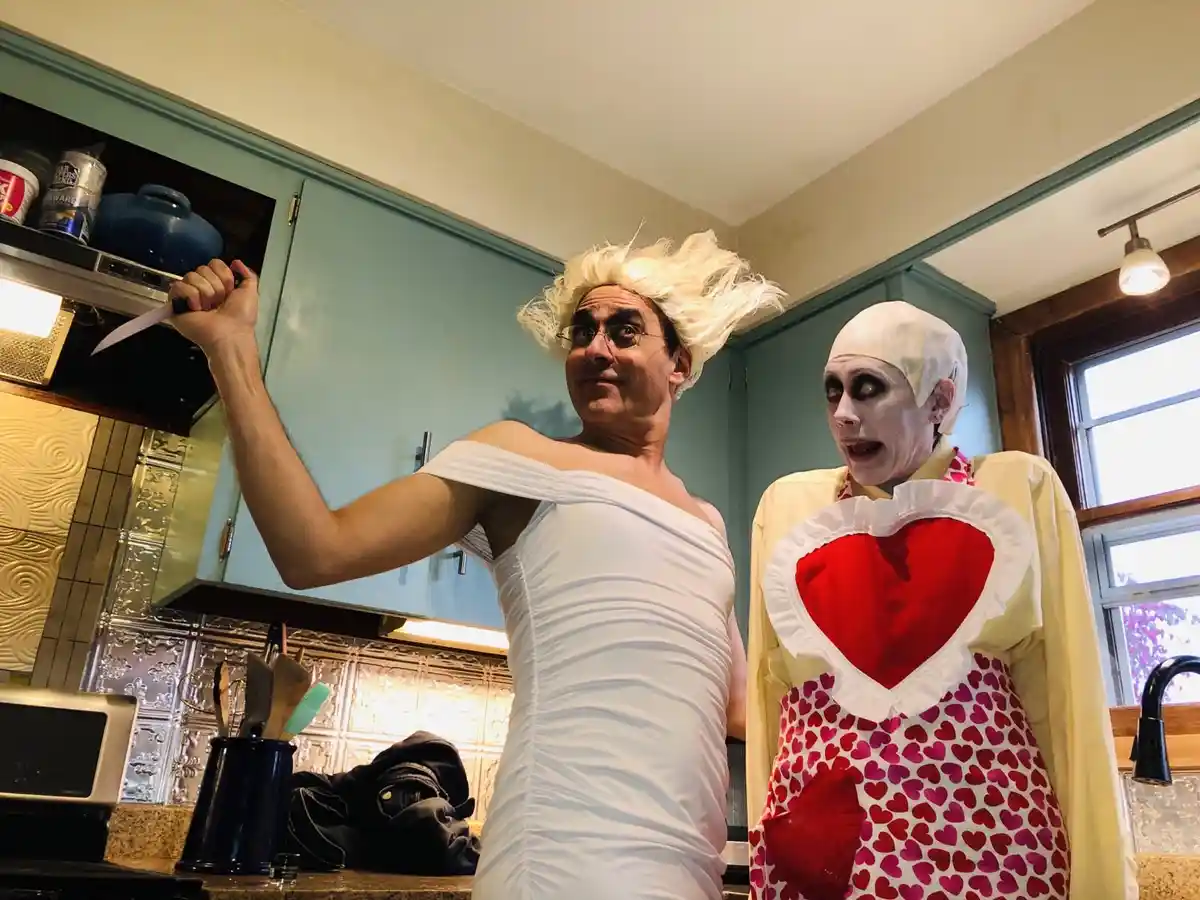 Two people in a kitchen, one dressed as a man in a white toga holding a knife, the other dressed as a pale-faced chef with a…