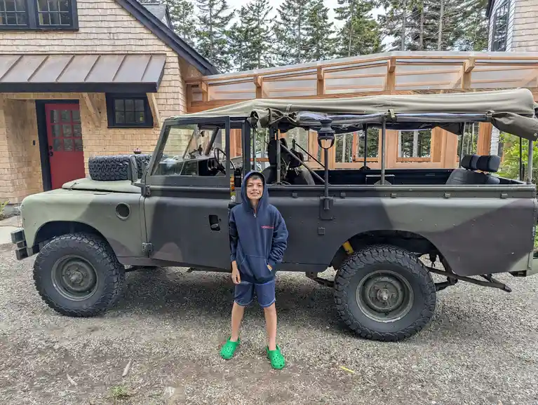 A young person wearing a blue hoodie, blue shorts, and bright green Crocs stands confidently in front of a large, rugged…