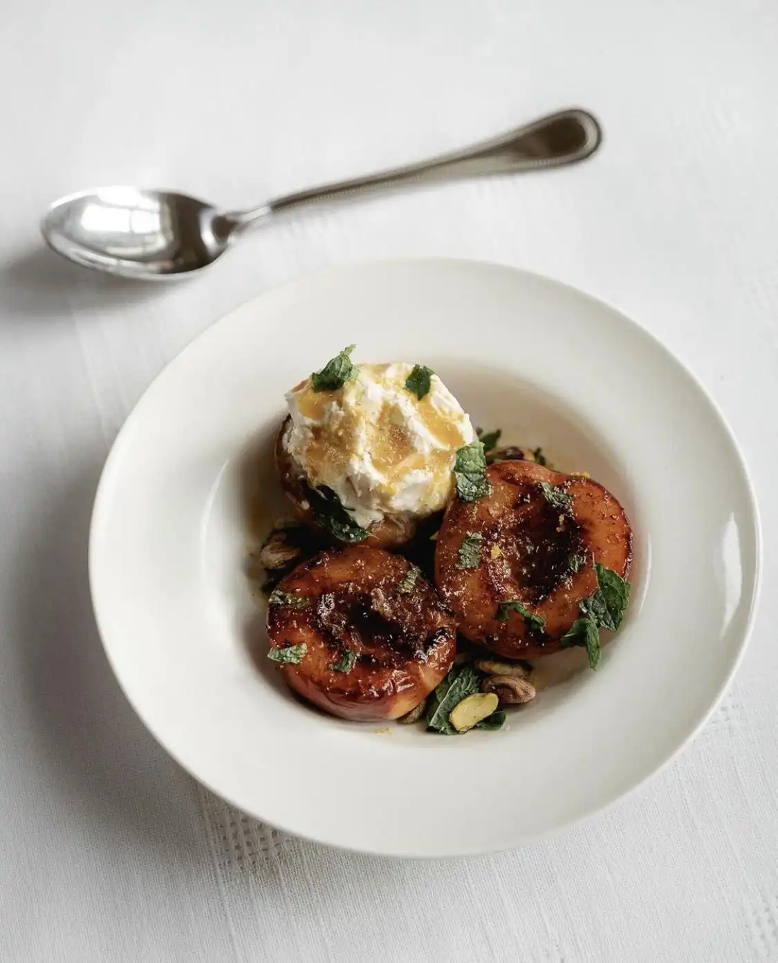 A white bowl holds caramelized peach halves topped with a scoop of vanilla ice cream, garnished with mint and pistachios…