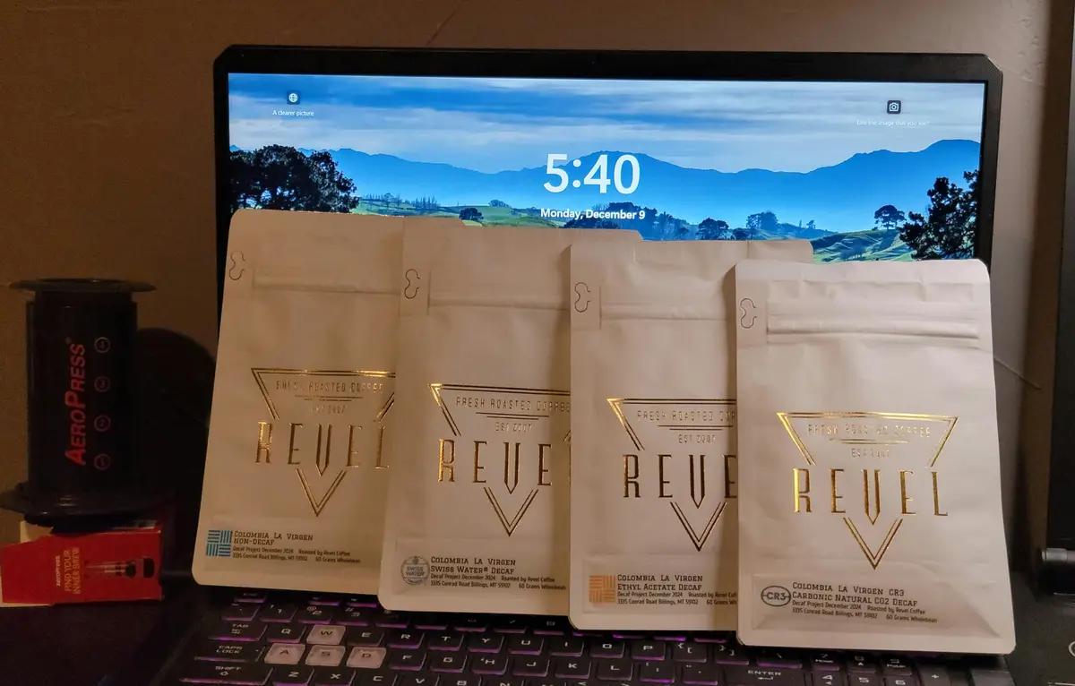 Four white bags of Revel Coffee decaf varieties are arranged in a row on a laptop keyboard, with the screen behind them…