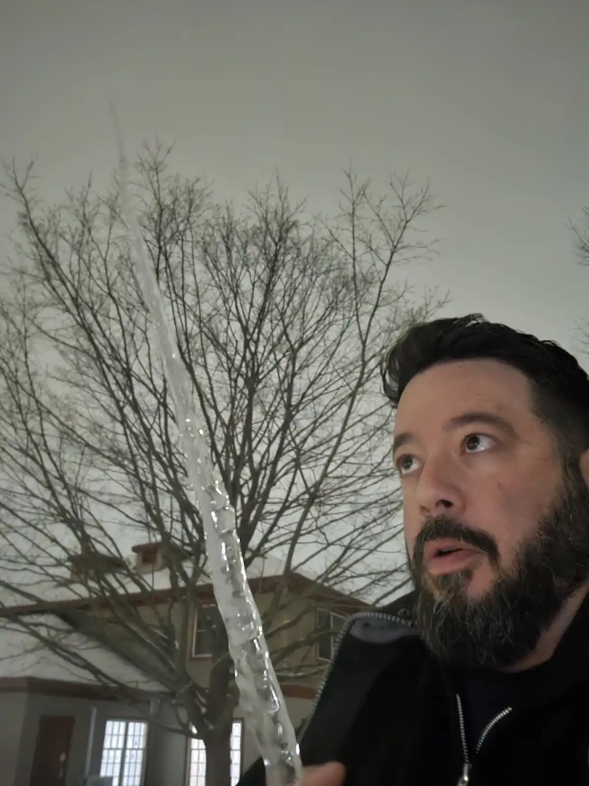 A man with a beard and dark hair, wearing a black jacket, looks upward in surprise as a large, clear icicle hangs vertically…