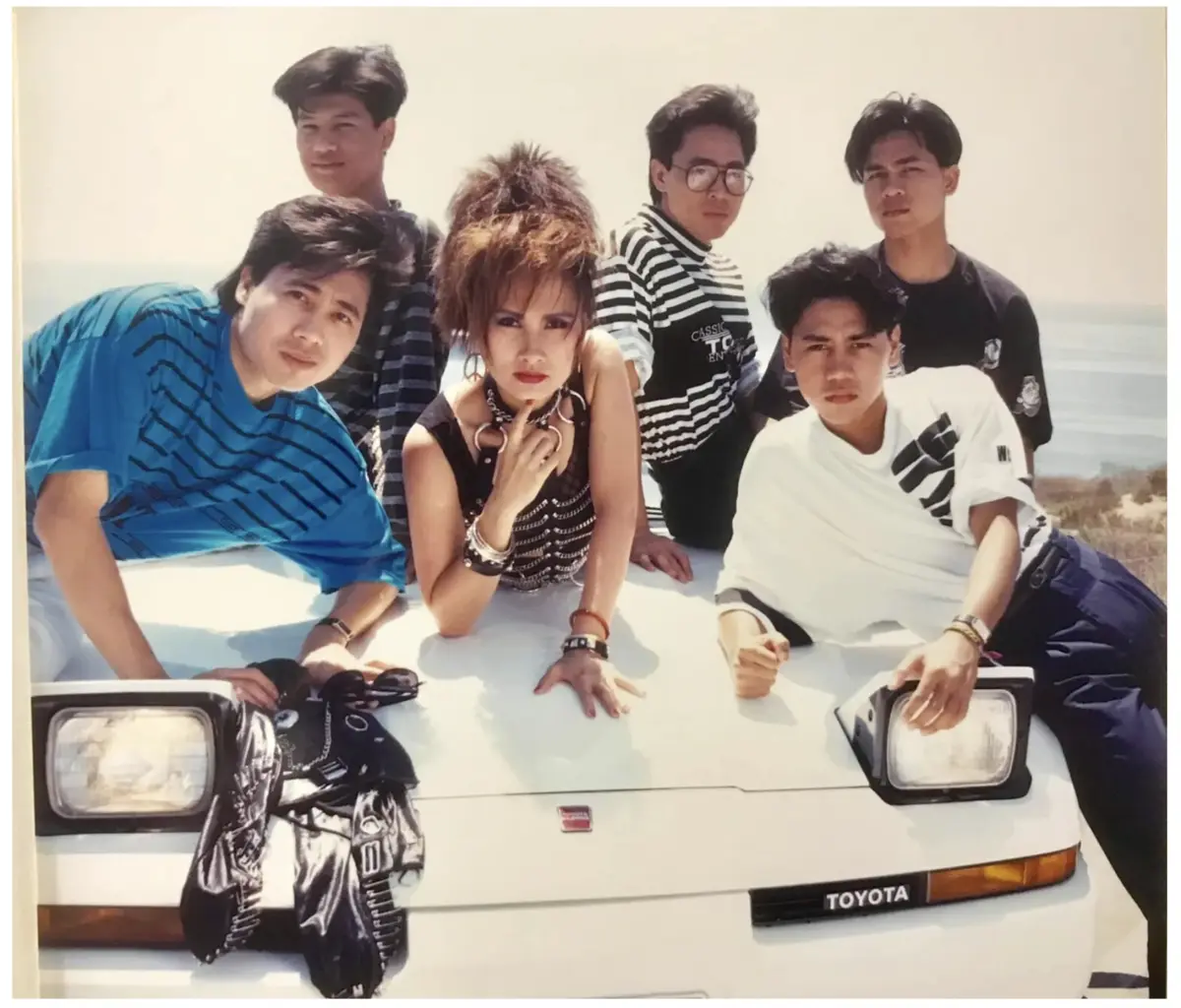 A vintage 1980s photo shows six people posing on the hood of a white Toyota car against a bright, hazy background. The group…