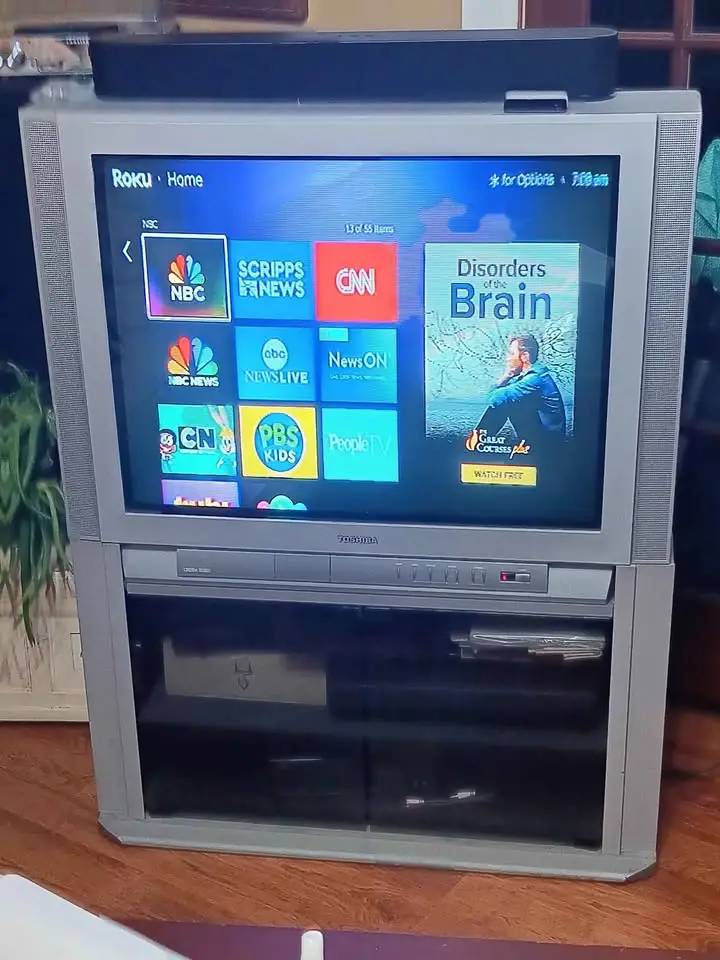 A silver Toshiba CRT television with a built-in DVD player is displaying a Roku home screen with various news channel apps…