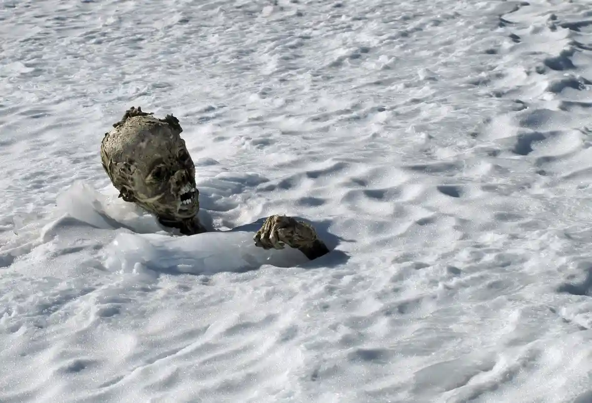 A weathered, grimacing human skull and one gnarled hand emerge from a snowy, icy landscape, with the overlaid text reading…