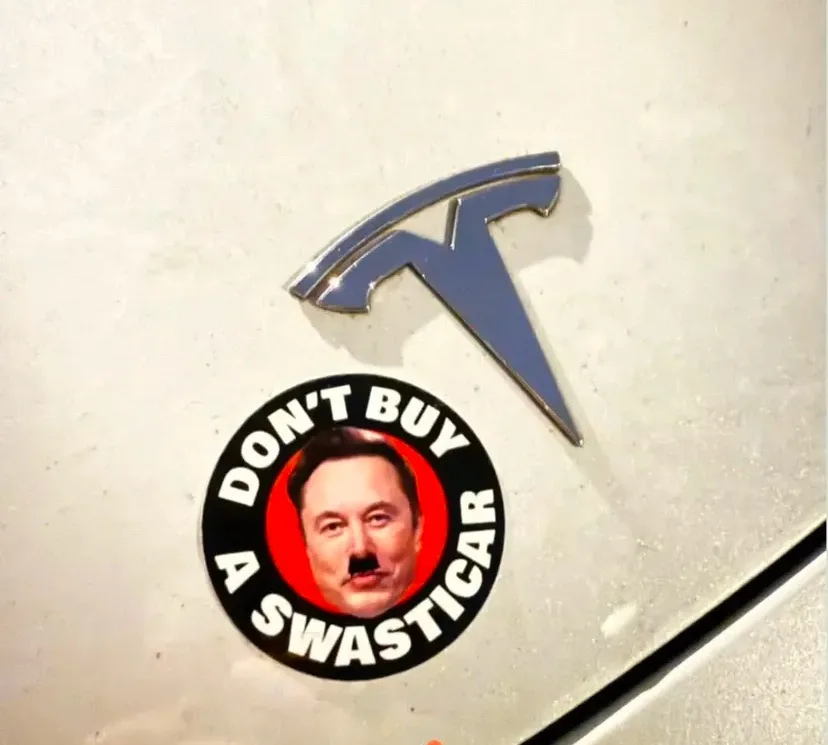 A close-up photo shows a Tesla car emblem on a white vehicle, with a circular sticker directly below it. The sticker…
