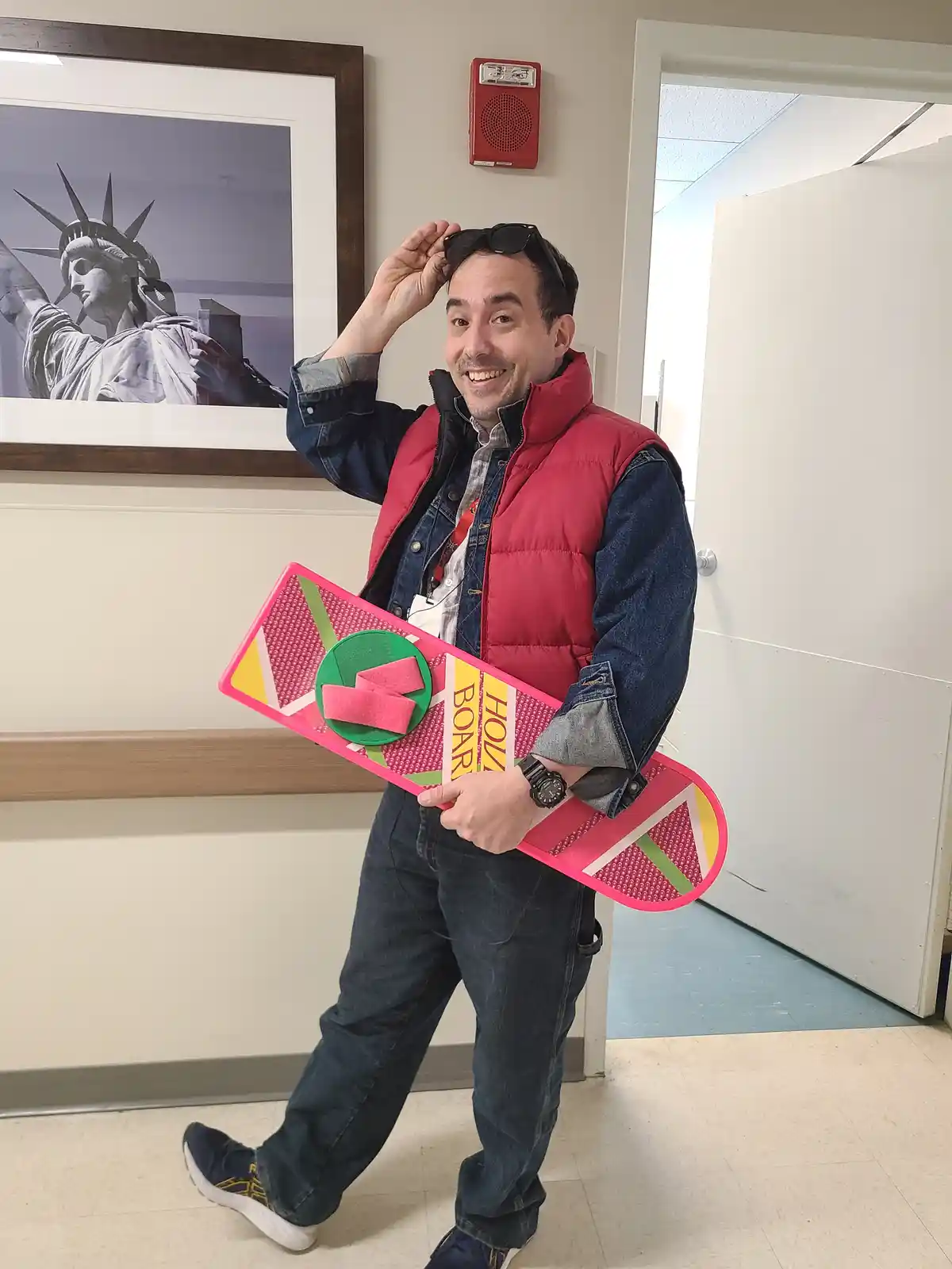 A man in a red vest and denim jacket poses indoors holding a pink, geometrically patterned hoverboard prop with 'HOLLYWOOD…