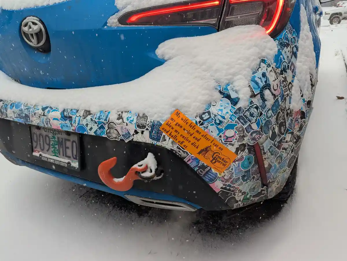 A blue Toyota car covered in snow, with its rear bumper and side heavily adorned with numerous stickers. An orange Cracker…
