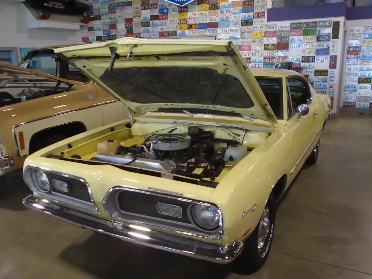 A pale yellow classic Plymouth Barracuda with its hood open, revealing the engine, is parked indoors in front of a wall…