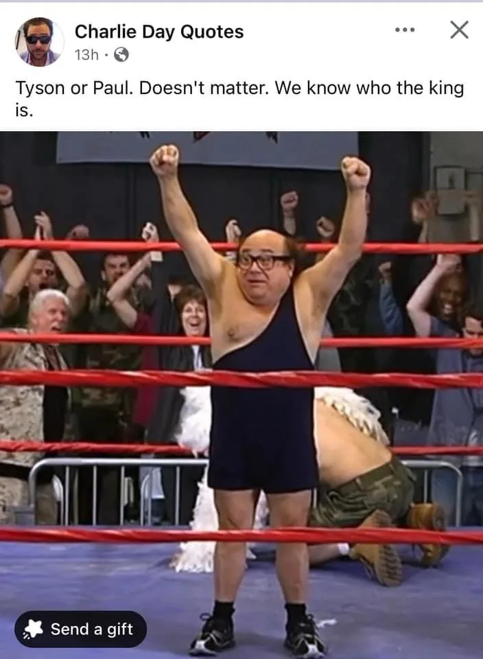 A man in a dark blue wrestling singlet and glasses stands triumphantly in a boxing ring with both fists raised, celebrating…