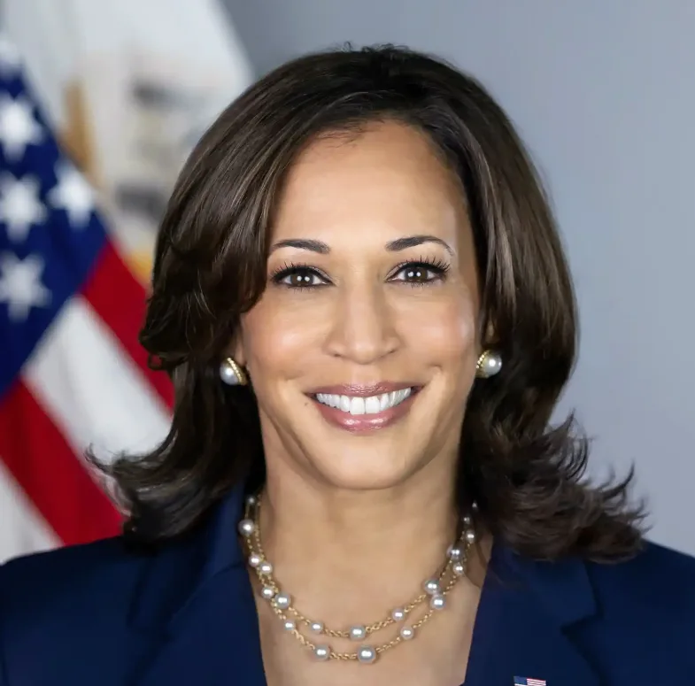 a formal portrait of Kamala Harris smiling, wearing a dark blue suit, pearl necklace, and pearl earrings, with an American…