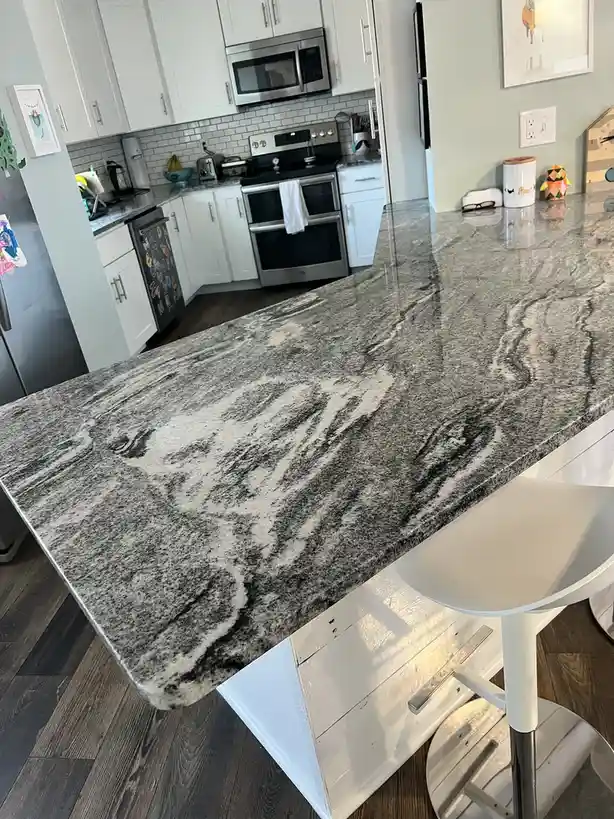 A photo of a modern kitchen with a large granite countertop that has dark and light swirling patterns resembling a tiger's…